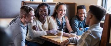 Five young people sit at a table in a restaurant, there are two coffee cups on the table. 