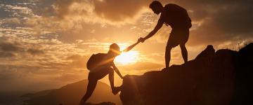 silhouette of two people hiking while one offers a hand to help pull the other up the hill