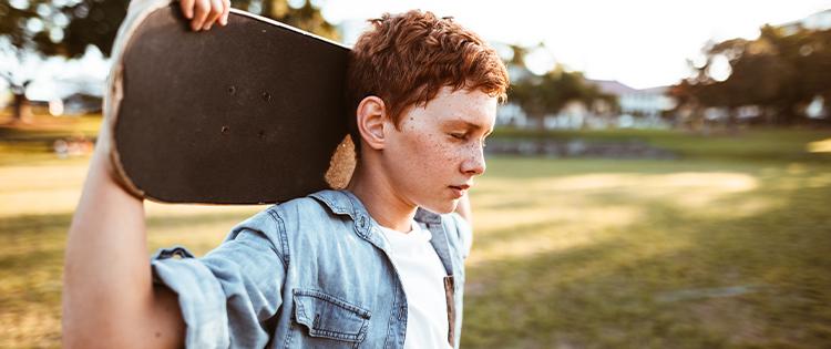 A Young man stands in a field holding a skateboard behind his head.