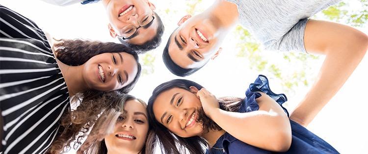 Five young people stand in a huddle and they look down into the camera in the middle.