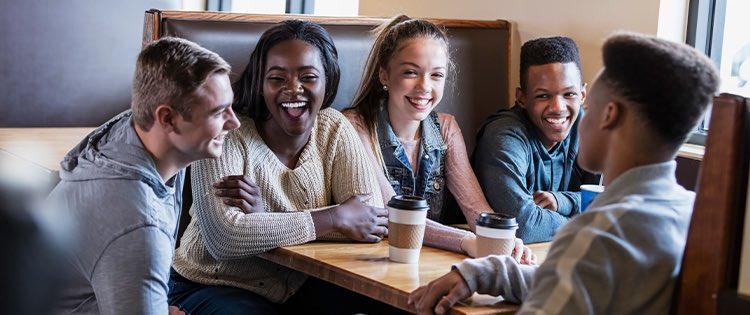 Five young people sit at a table in a restaurant, there are two coffee cups on the table. 