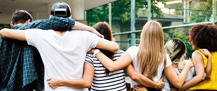 Five people stand with arms linked behind their backs and over their shoulders. The look off into the distance.