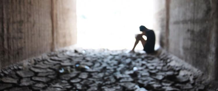 Silhouette of teen sitting on ground against a wall with head in hands.