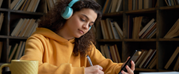 Teenage girl in yellow sweatshirt and blue headphones is holding her phone and writing things down.